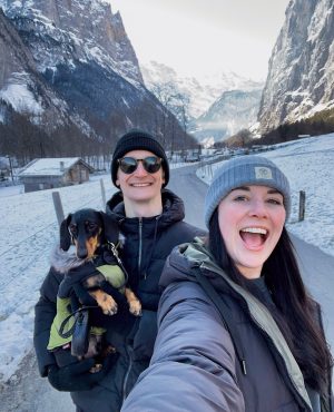 woman and man holding small dog smile with snowy swiss village behind them